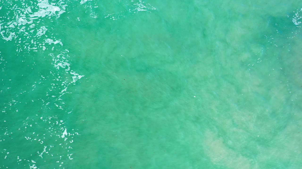 Overhead view of green sea water in Los Cabos, Mexico, with white cresting water along the left edge and pale sandy shallows visible below, no visible people.