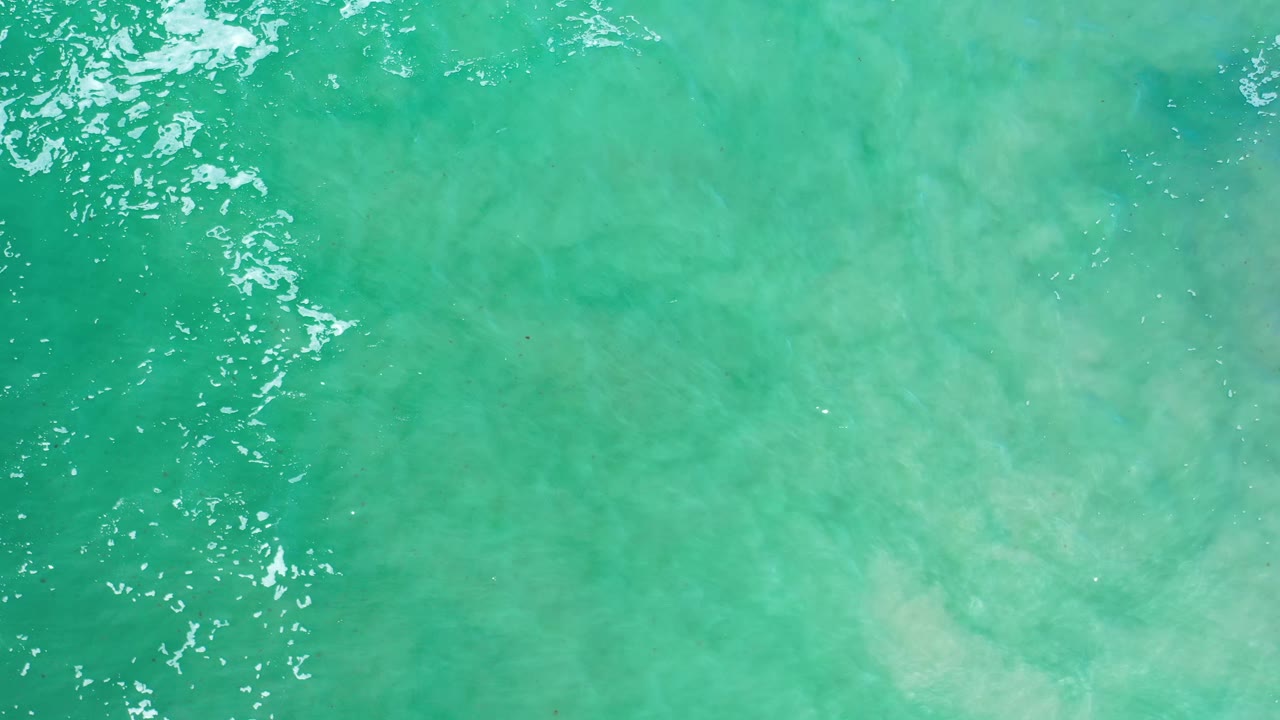 Turquoise-green sea surface in Los Cabos, Mexico, with a bright patch of white cresting water near the upper left and cloudy sandy shallows beneath, no visible people.