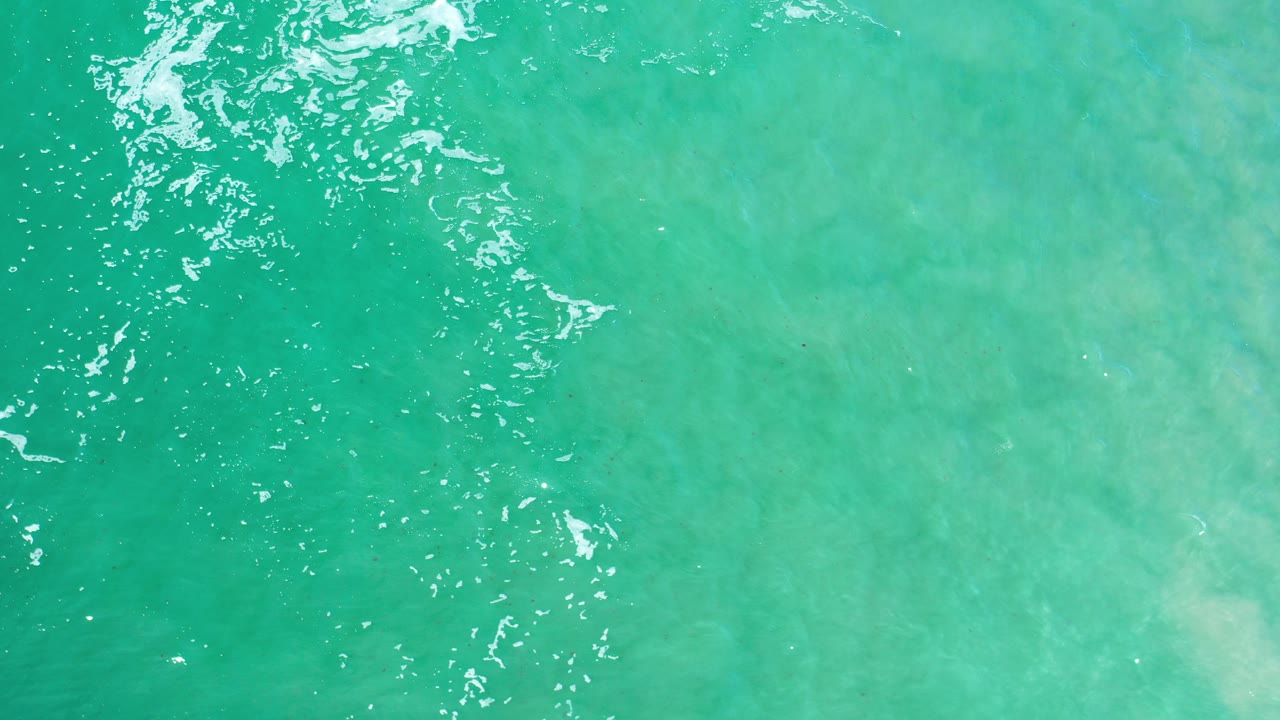 Opening frame of Los Cabos turquoise ocean seen from above, with white ripples near the upper left and no visible people.