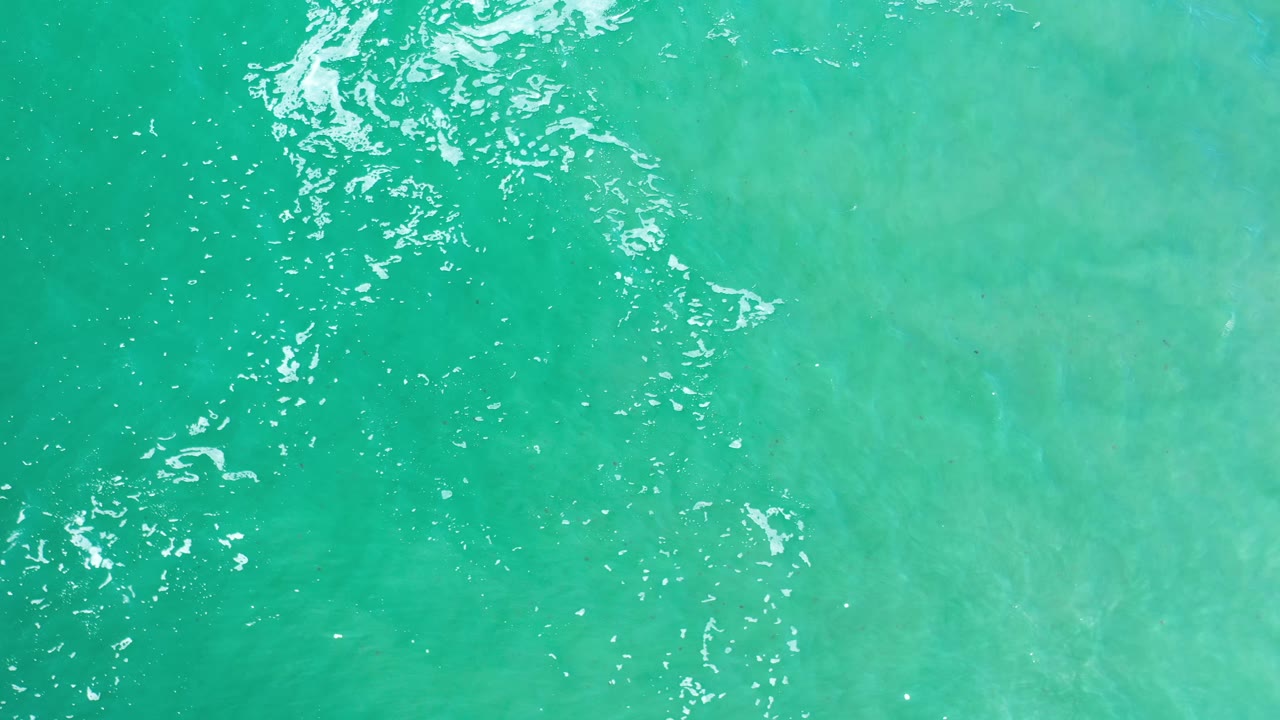 Aerial frame over Los Cabos ocean water, showing a broader patch of pale underwater color and scattered white highlights, with no visible people.