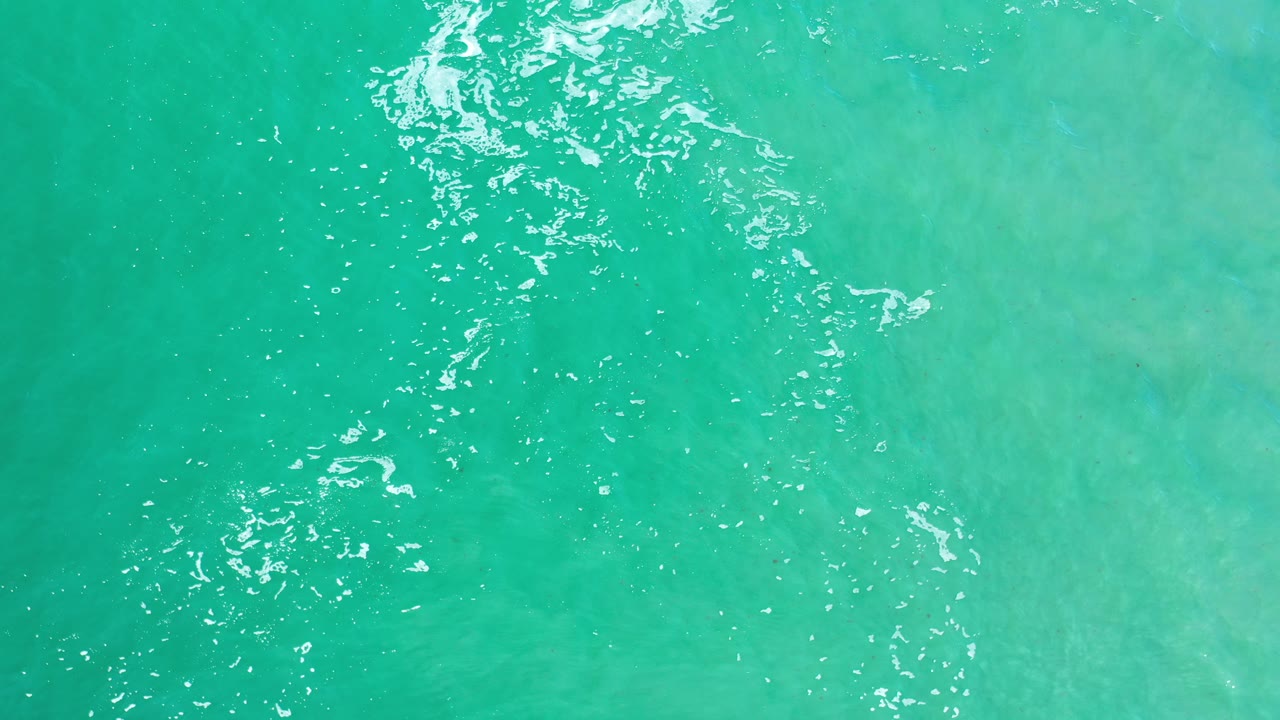 Aerial turquoise water with white shimmer and soft pale areas beneath.