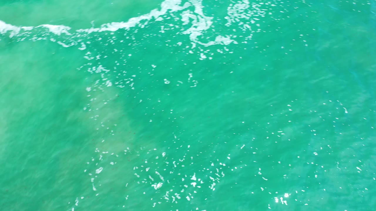 Overhead Los Cabos ocean frame with clear green-blue water, scattered sun sparkles, faint foam along the top, and no visible people.