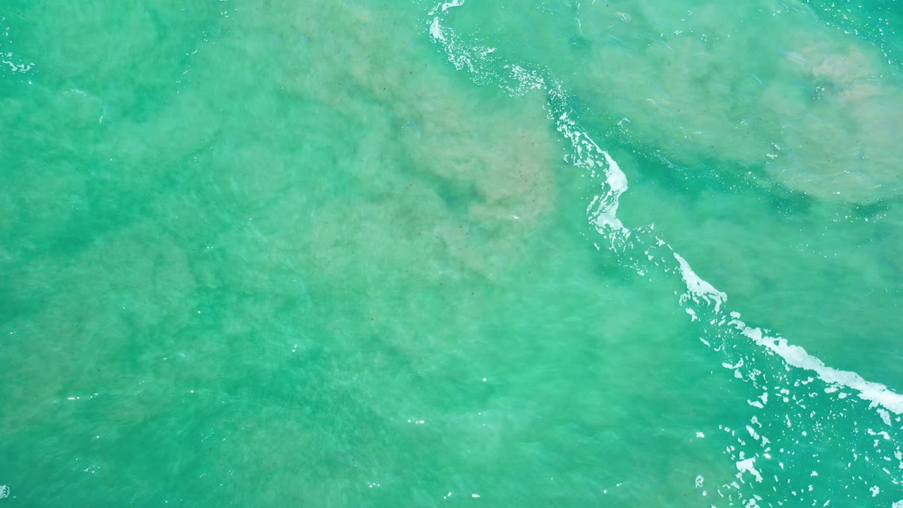 Overhead view of clear turquoise water in Los Cabos, Mexico, with pale seabed detail and a narrow broken foam line, no visible people.