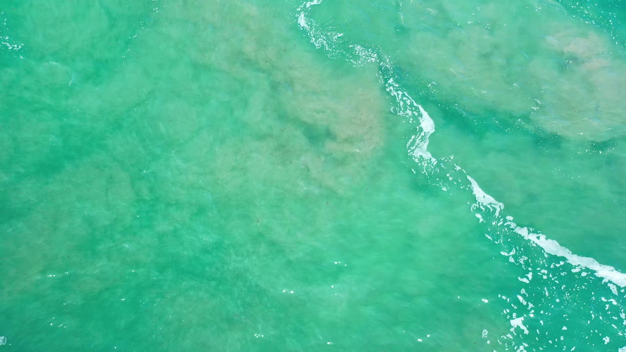 Clear green-blue sea from above in Los Cabos, Mexico, with a diagonal streak of white foam crossing visible shallow-bottom texture, no visible people.