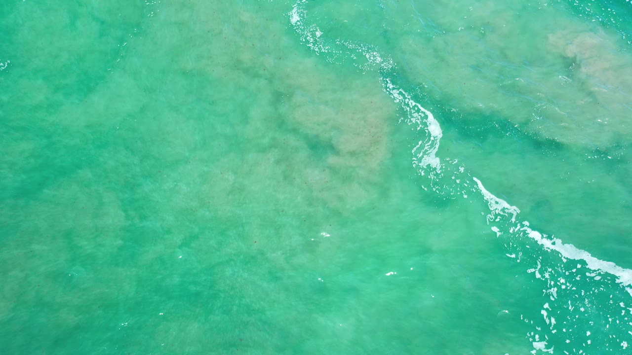 Overhead view of green seawater in Los Cabos, Mexico, with pale sandy bottom features, a diagonal white foam line near center, and no visible people.