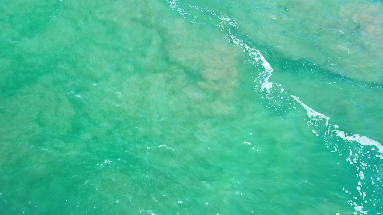 Overhead view of turquoise ocean water in Los Cabos, Mexico, with pale submerged sand patches, a diagonal white foam streak, and no visible people.