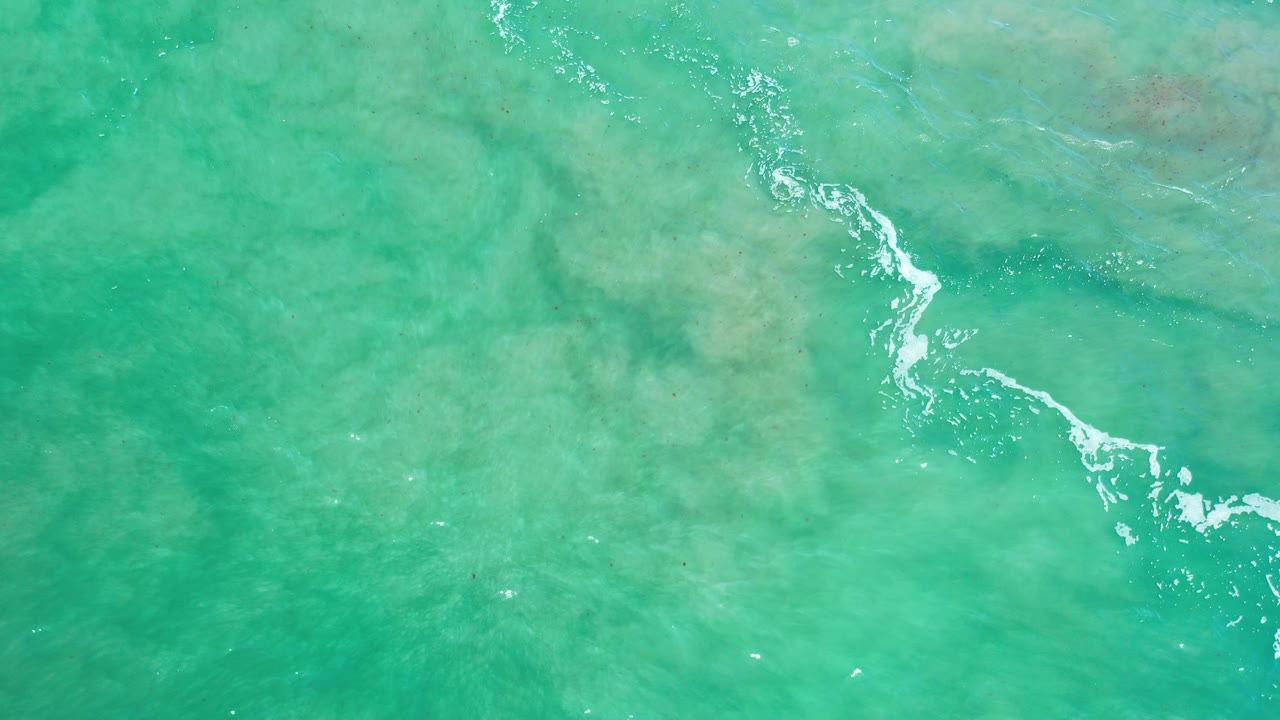 Overhead view of sea green water in Los Cabos, Mexico, with a white foam streak crossing shallow reef patches and no visible people.