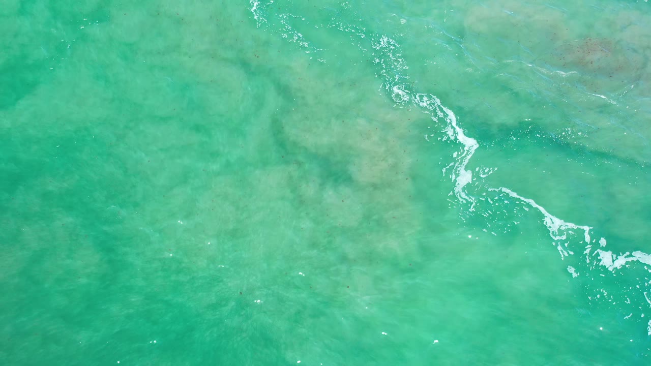 Sea green water in Los Cabos, Mexico, shows a diagonal line of white foam drifting over muted reef shapes with no visible people.
