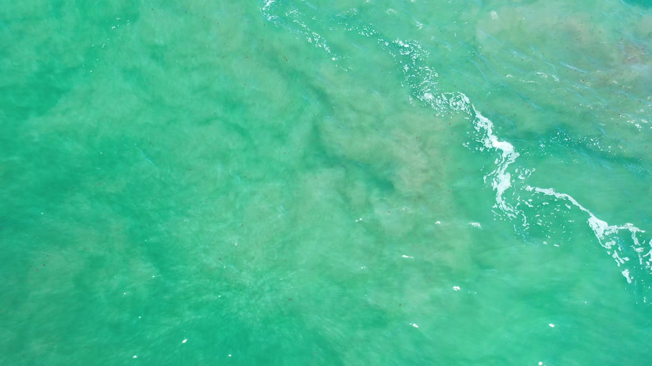 Overhead view of turquoise ocean water in Los Cabos, Mexico, with pale submerged texture and a small diagonal foam streak, no visible people.