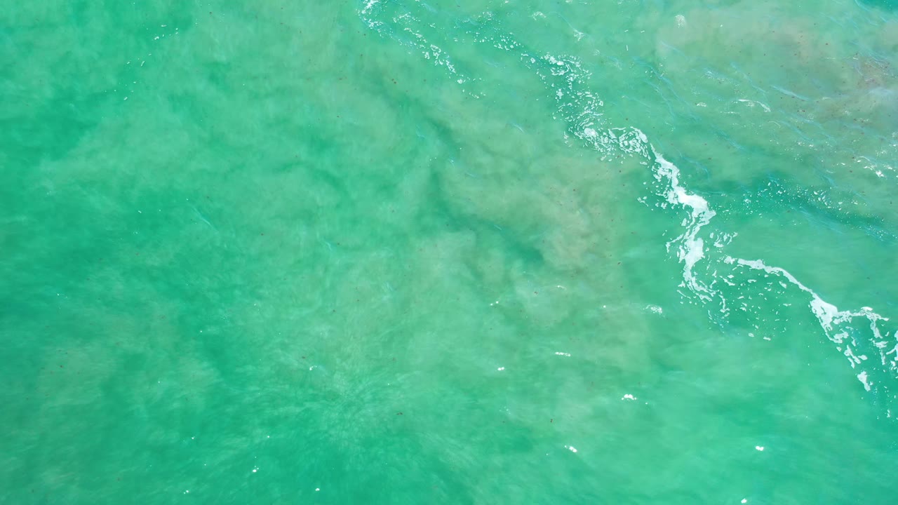Green-blue Los Cabos ocean surface seen from above, with a thin white ripple line near the center and soft submerged shapes below, no visible people.