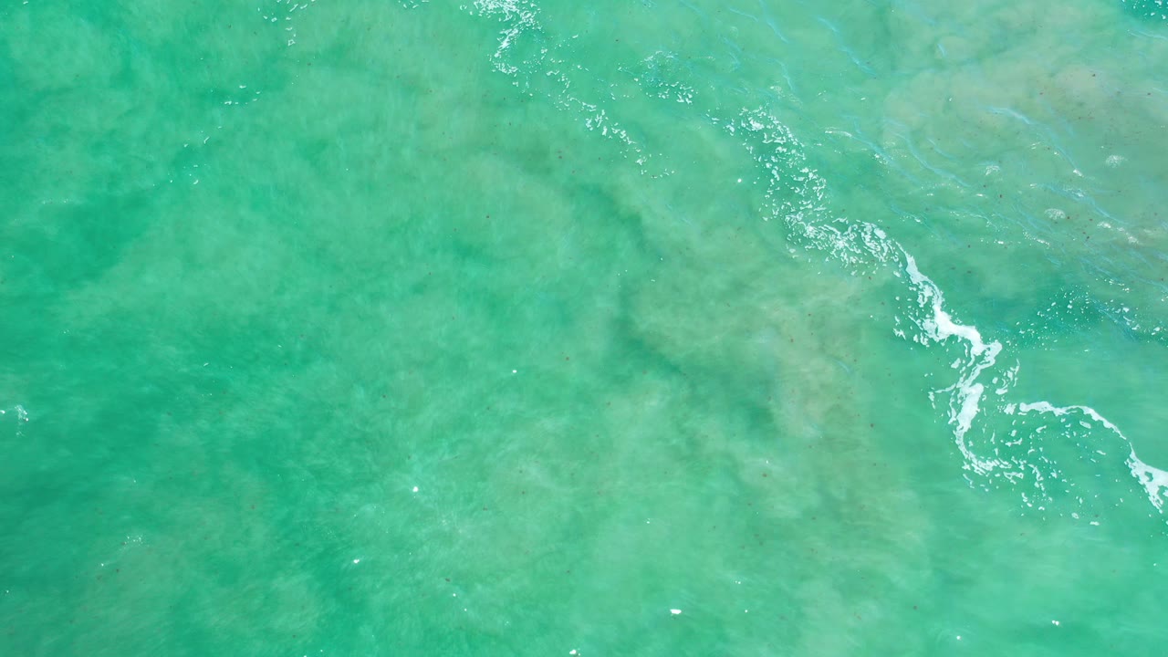 Overhead view of turquoise-green water in Los Cabos, Mexico, with a narrow white foam streak crossing pale submerged rock or sand patterns and no visible people.