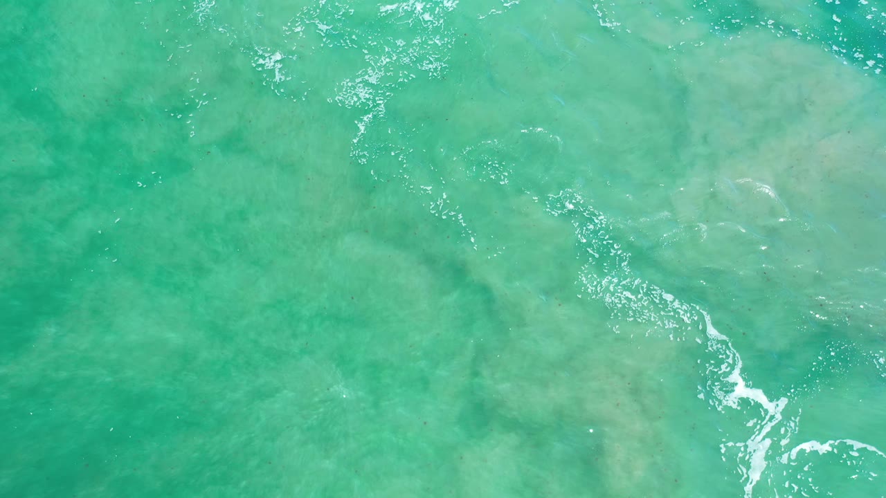 Aqua water in Los Cabos shows a diagonal streak of white foam crossing over muted rocky shallows, with no visible people.