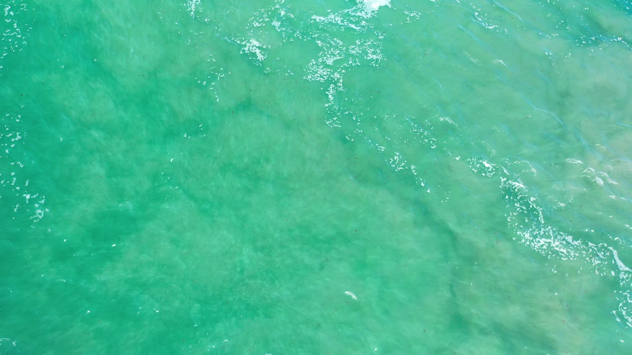 Overhead view of turquoise ocean water in Los Cabos, Mexico, with pale sand visible below the surface, scattered white ripples, and no visible people.