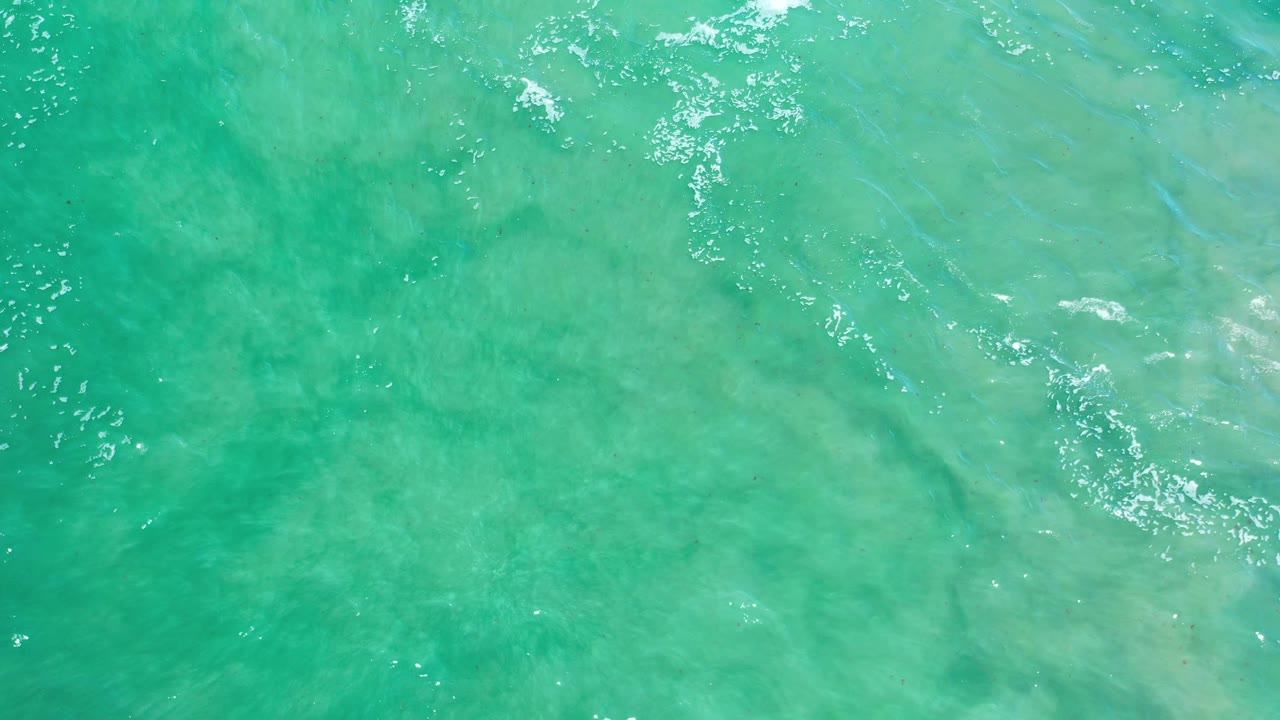 Aerial ocean surface with turquoise water, soft sand color, and ripple clusters.