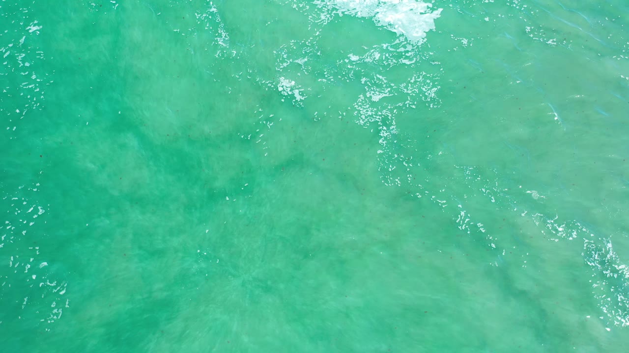 Overhead Los Cabos ocean frame with aqua water, a white foam patch near the upper edge, and no visible people.