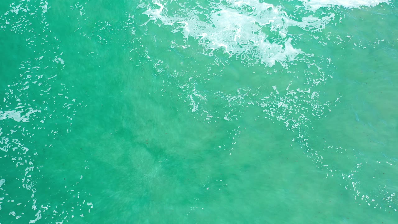 Overhead blue-green ocean surface in Los Cabos, Mexico, with a wider foam patch near the top and no visible people.