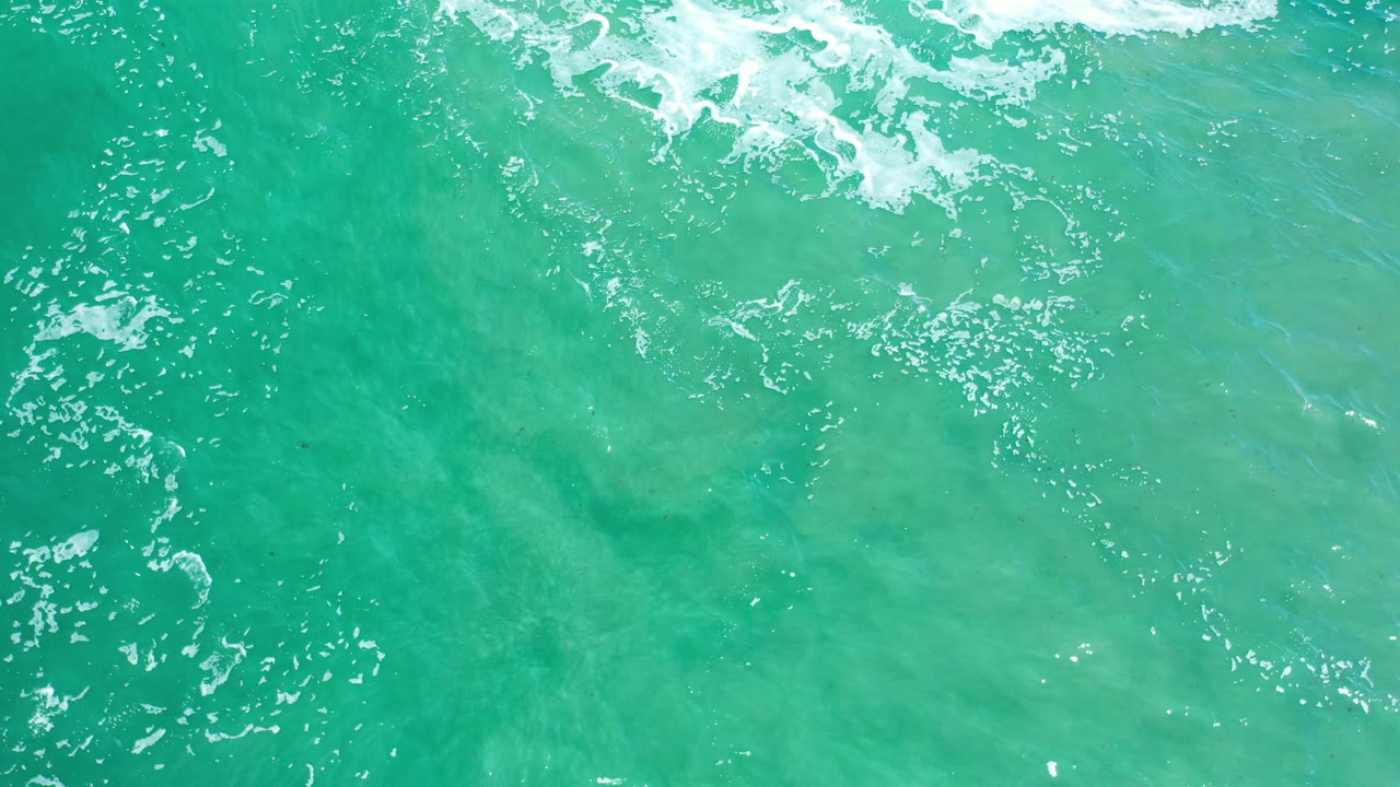 Turquoise green ocean surface in Los Cabos, Mexico, seen from above with white foam breaking across the upper frame and no visible people.