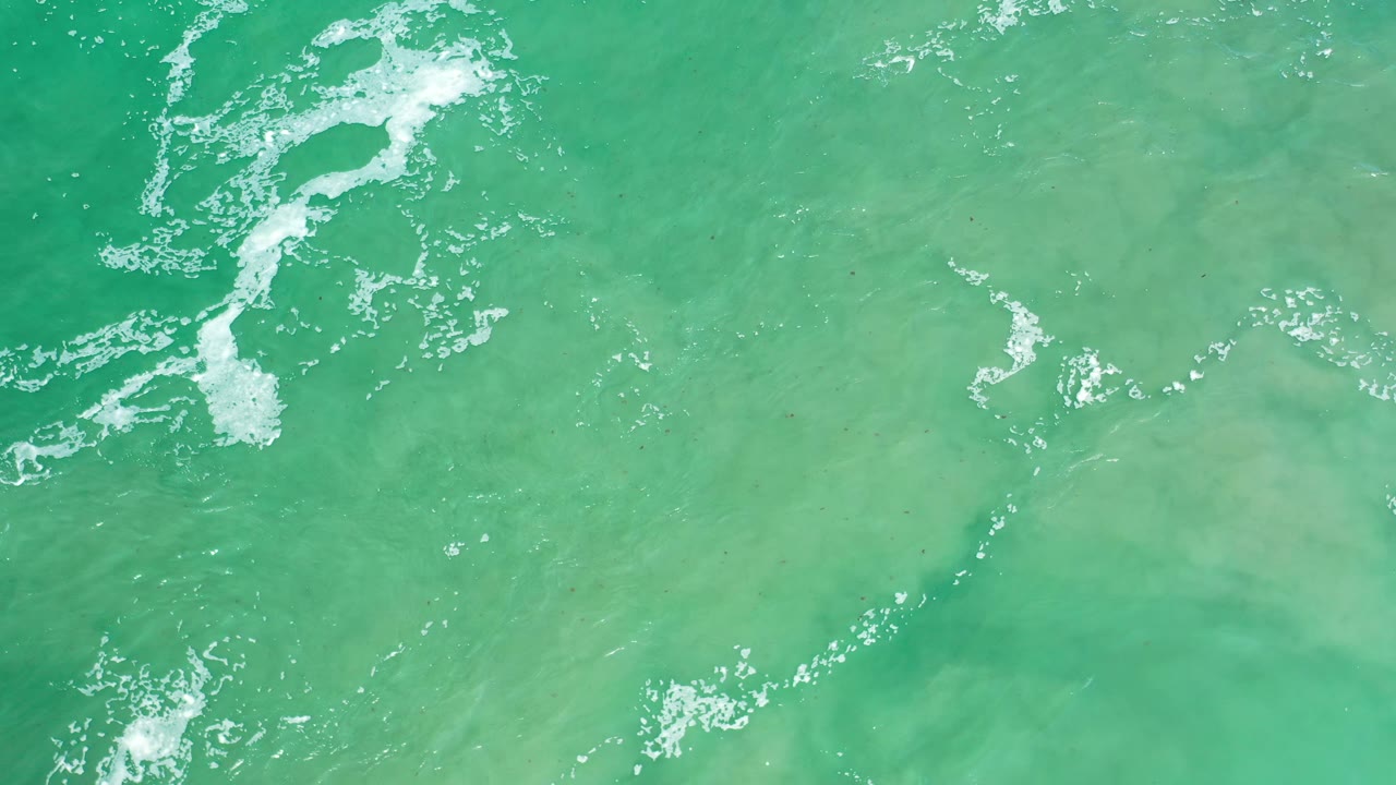 Overhead Los Cabos ocean frame with turquoise water, pale sandy shapes beneath the surface, scattered foam, and no visible people.