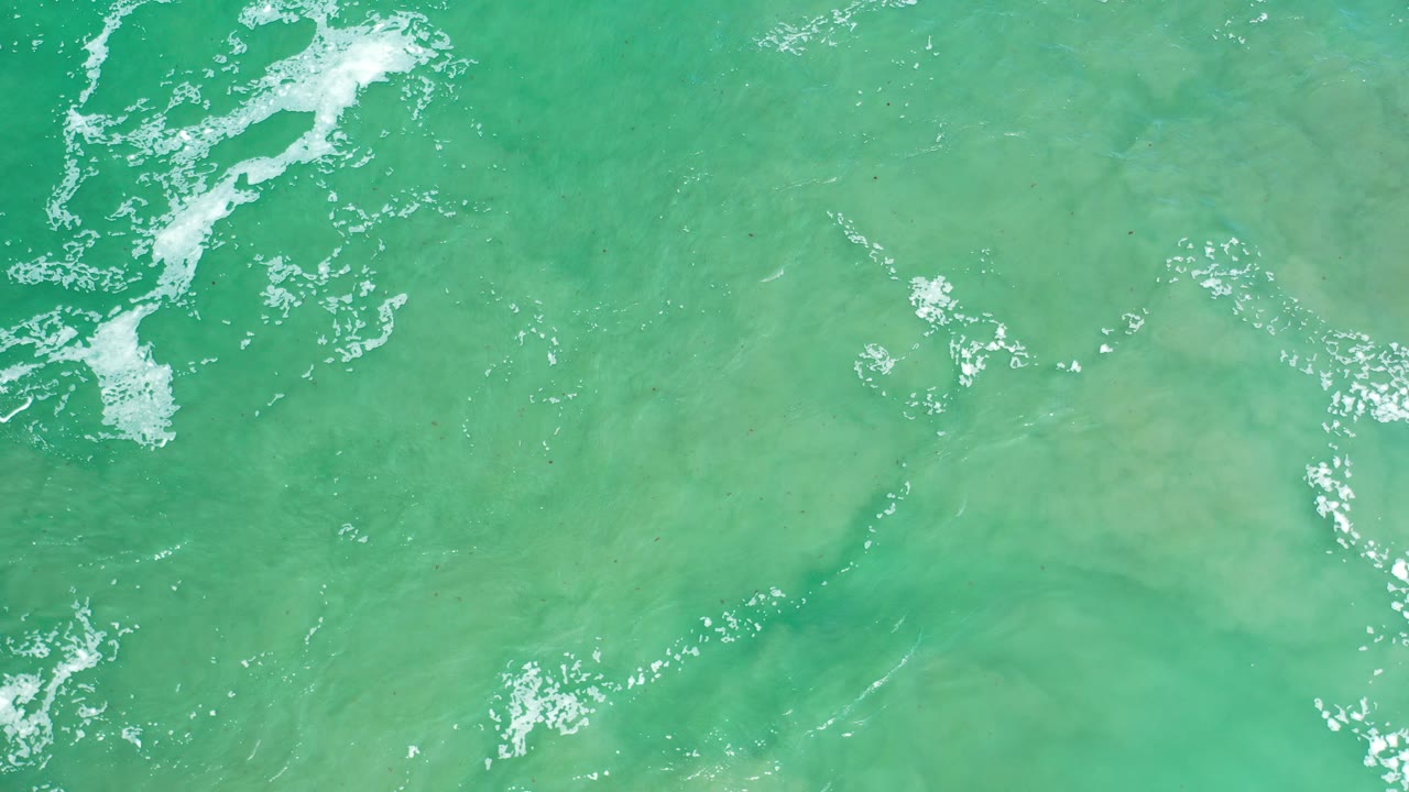Overhead view of green-blue ocean water in Los Cabos, Mexico, with white foam gathered near the upper left and no visible people.