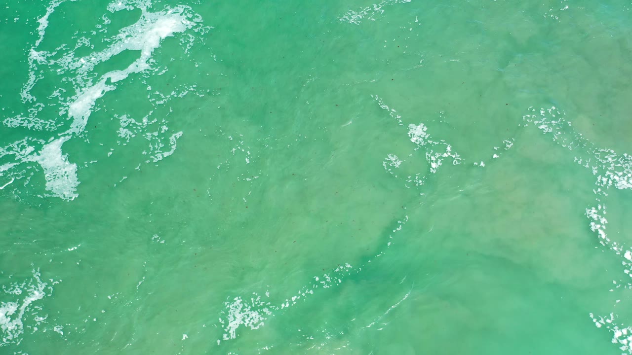 Overhead Los Cabos ocean frame with turquoise water, faint sandy-looking patches below the surface, scattered foam, and no visible people.