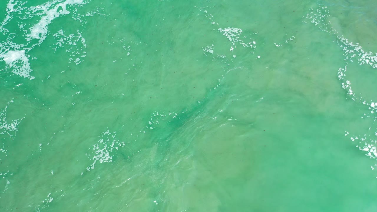 Overhead view of turquoise-green sea water in Los Cabos, Mexico, with scattered white foam at the upper edge and no visible people.