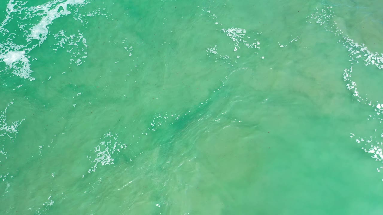 Aerial-looking view of Los Cabos sea water with pale underwater patches, small foam clusters, and no visible people.