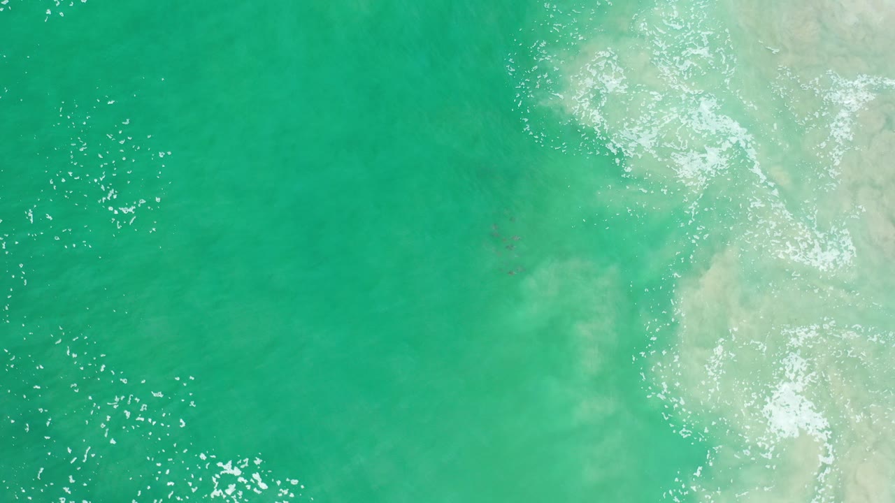 Overhead view of green ocean water in Los Cabos, Mexico, with scattered sun glints, pale foam along the right edge, and no visible people.