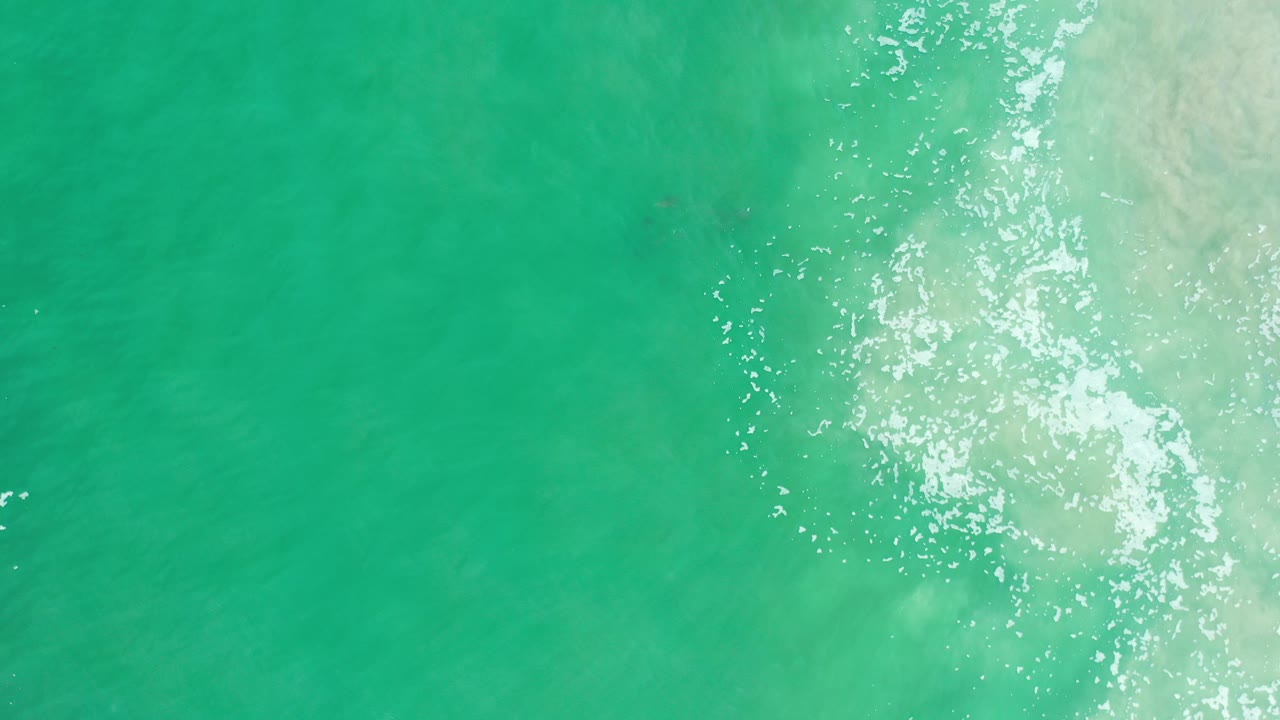 Overhead view of aquamarine water in Los Cabos, Mexico, with scattered white foam and bright sun glints along the right side, no visible people.