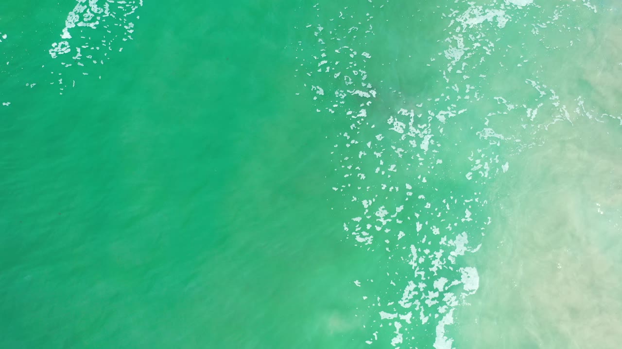 Clear green-blue ocean water in Los Cabos, Mexico, seen from above with bright foam streaks along the right side and no visible people.
