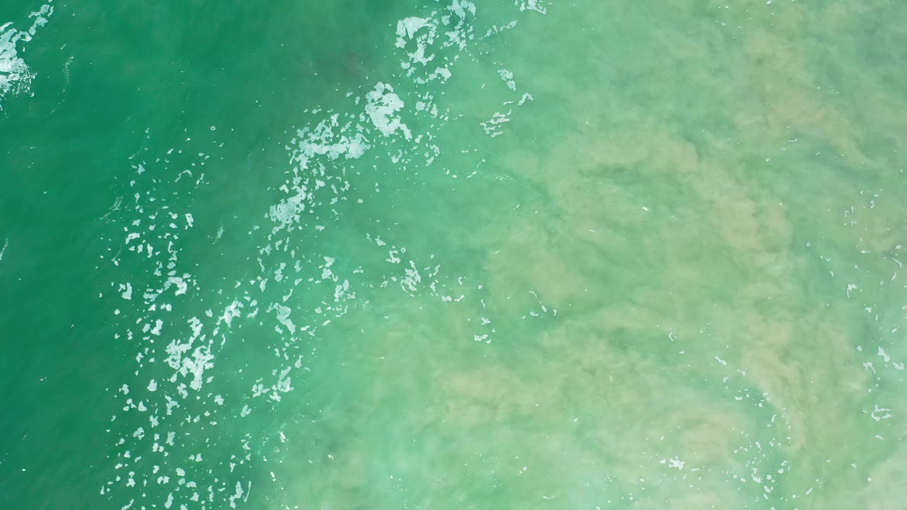 Overhead Los Cabos view of green-blue seawater, pale suspended sand or foam spreading through the right side, and no visible people.