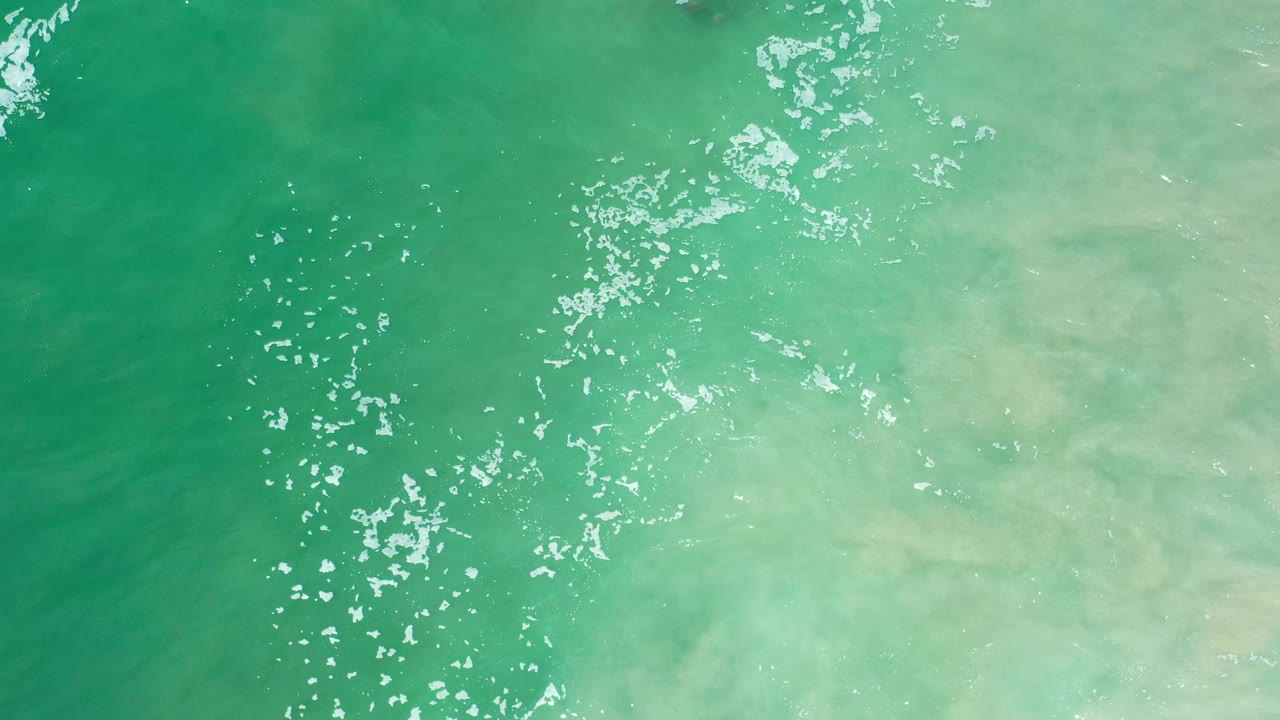 Overhead Los Cabos ocean frame with turquoise water, pale sandy coloration under the surface, a central scatter of white glints, and no visible people.