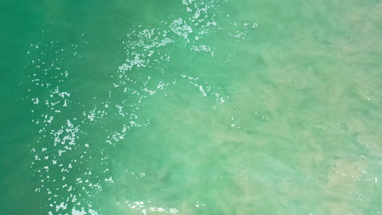 Overhead Los Cabos water frame with a brighter diagonal streak of sunlit ripples crossing green and pale aqua surface tones, with no visible people.
