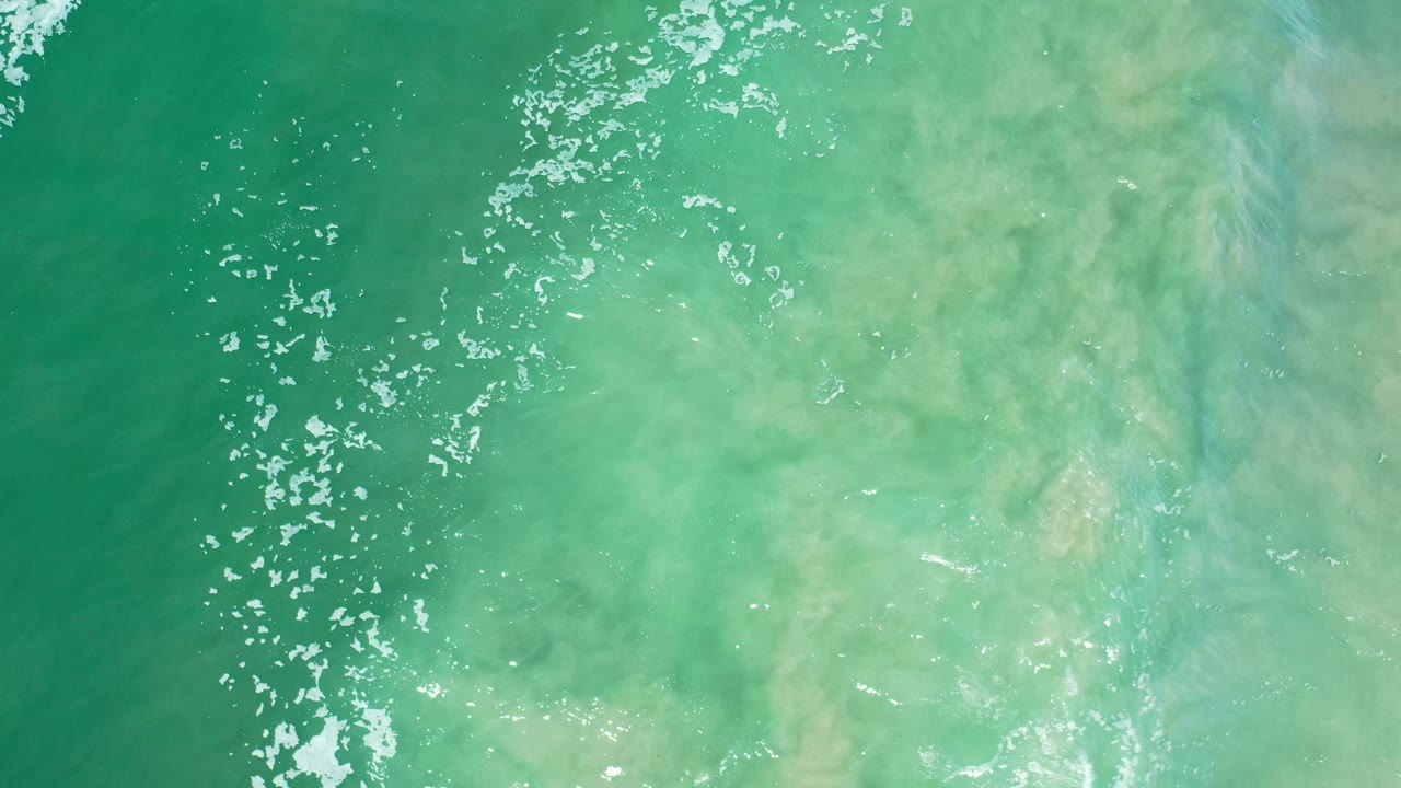 Blue-green Los Cabos water seen from above, with cloudy tan sand below the surface and a loose band of white foam along the left side, no visible people.