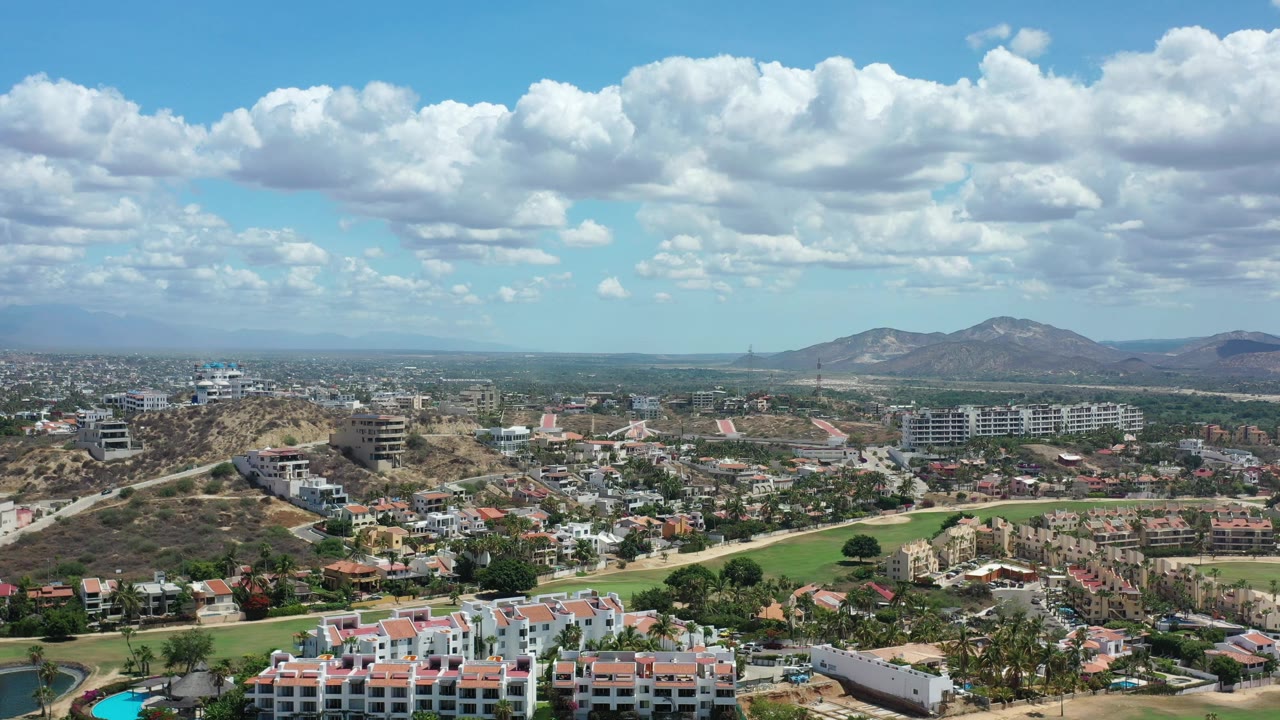 Opening aerial frame over Los Cabos, Mexico with hillside roads, residential buildings, distant mountains, large white clouds, and no visible people.