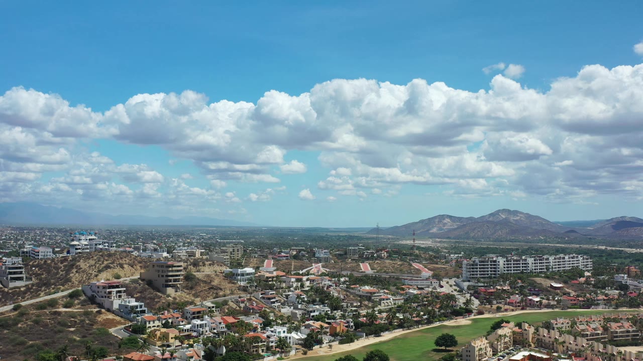 Los Cabos aerial frame with dry hills in the foreground, modern buildings across the midground, mountains on the horizon, and no visible people.