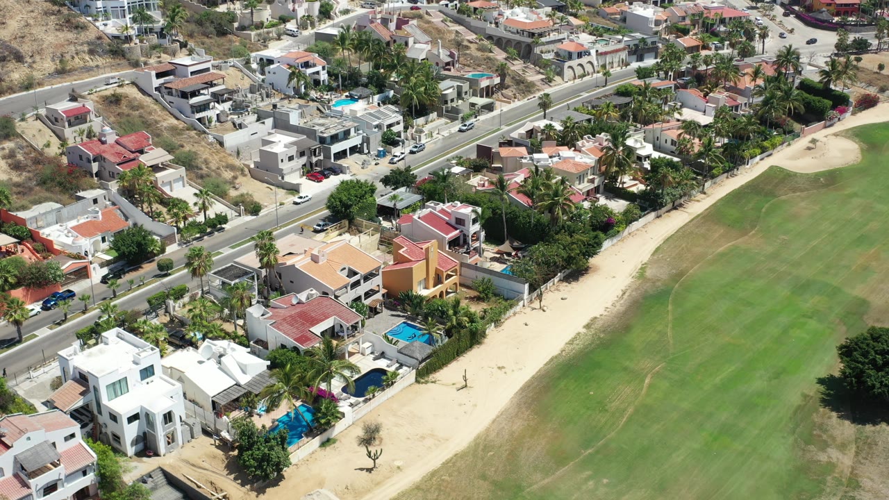 Aerial frame from Los Cabos, Mexico with red-roofed and white homes, blue backyard pools, cars along the road, palms, and the park grass occupying the right side with no visible people.