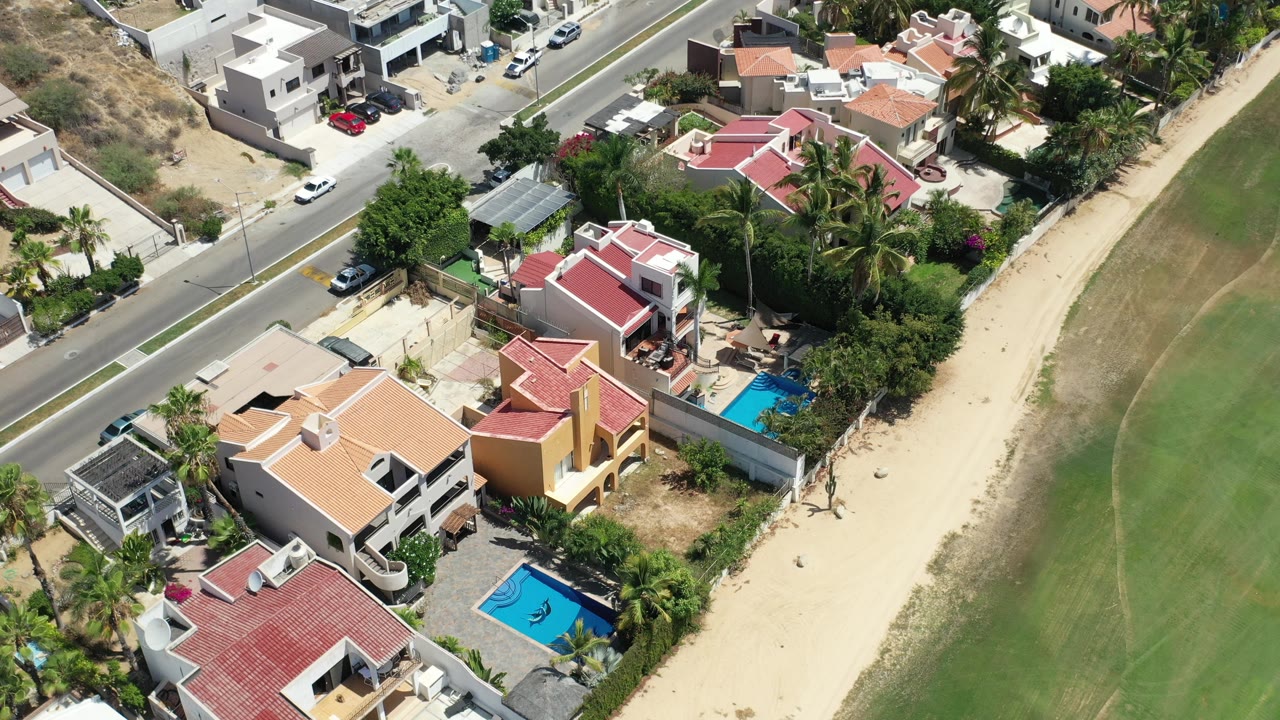 Aerial frame over Los Cabos, Mexico, showing tiled-roof houses, a paved road, sandy shoreline, green coastal strip, pools, palms, and no visible people.