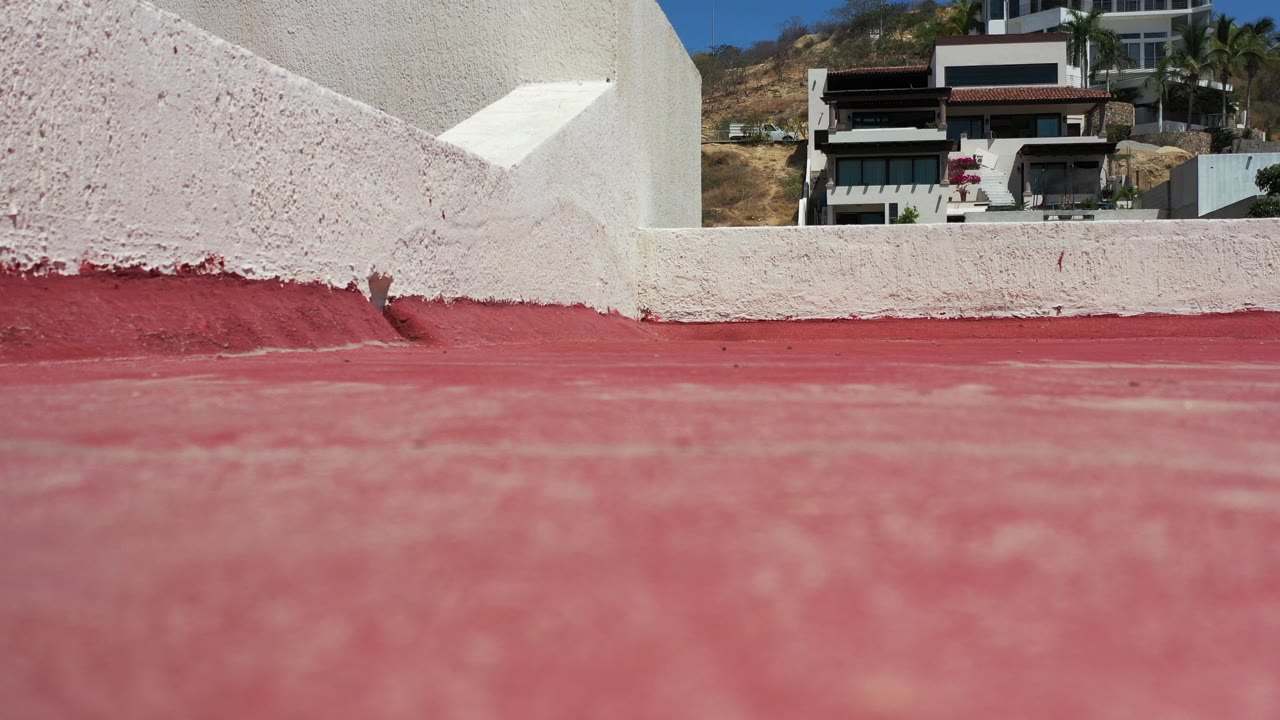 Low view across a red roof surface toward a white stucco wall and modern hillside home.