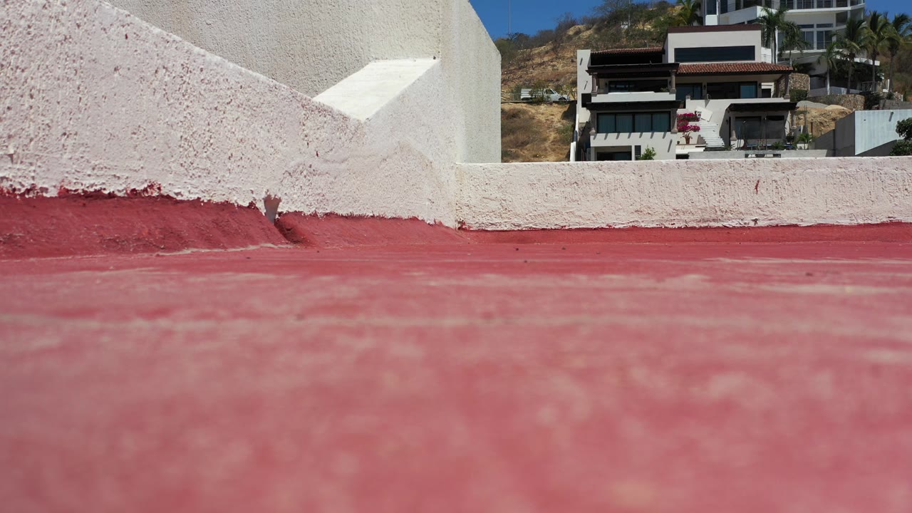 Red rooftop floor, white wall, and modern hillside building beyond.
