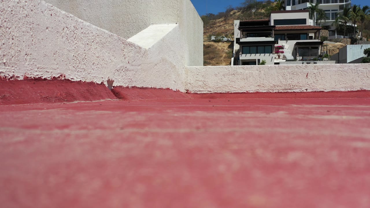 At 0:01, the Los Cabos rooftop remains framed low, with the red floor dominant, the white parapet edge sharp in the midground, distant houses, blue sky, and no visible people.