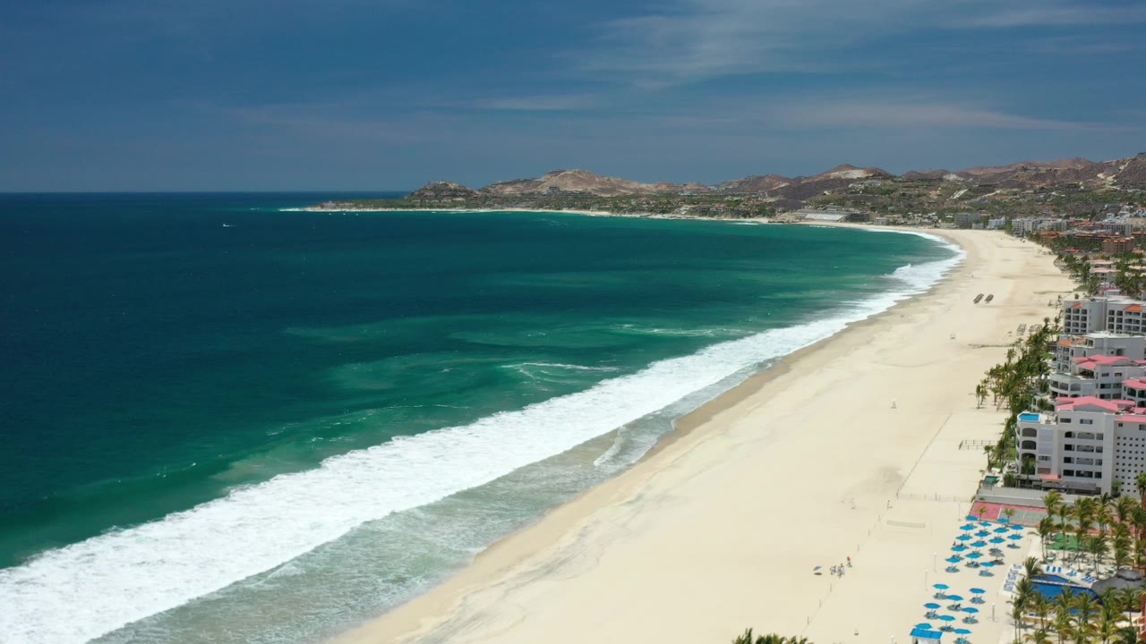 Aerial view of surf beside a wide sandy beach and resort buildings.