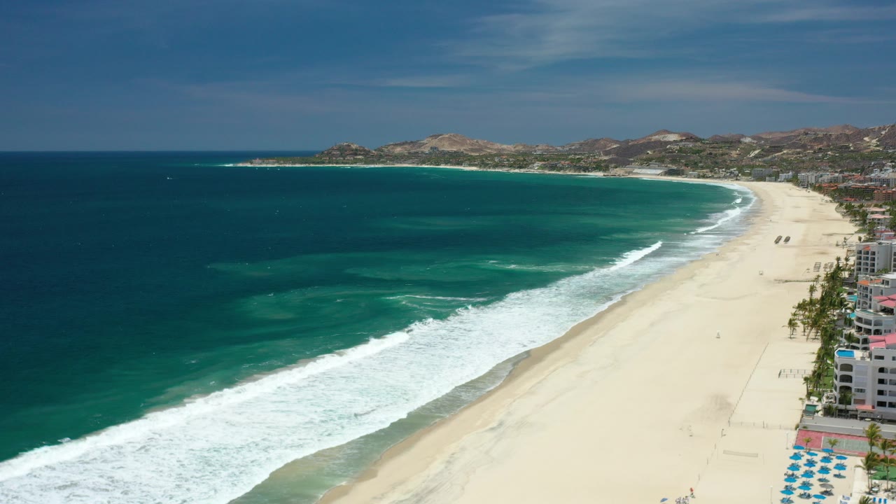 Opening aerial frame over a Los Cabos beach, with dark blue ocean at left, white surf on pale sand, low hills in the distance, and no visible people.