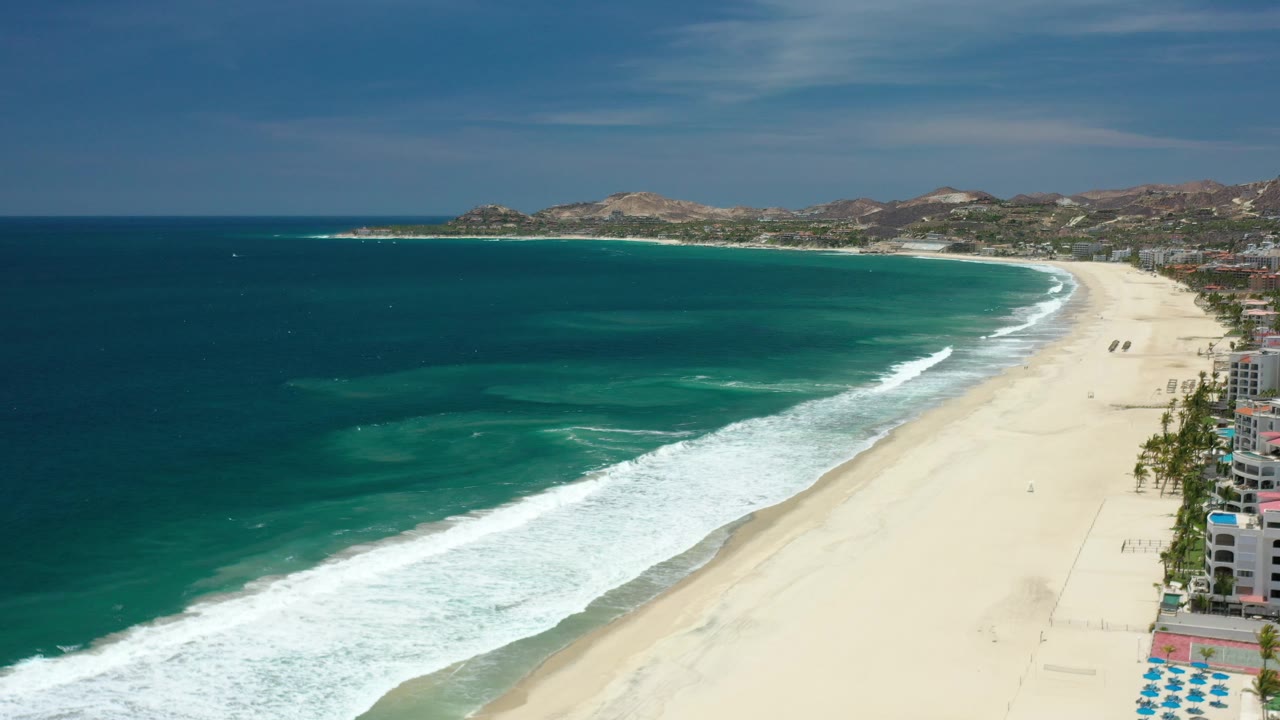 Elevated beach view with teal water, white waves, sand, and hills.