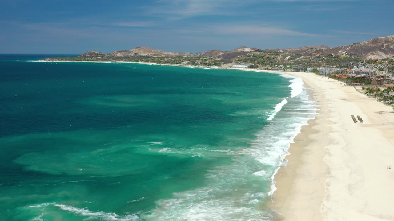 Aerial view over a Los Cabos, Mexico shoreline where aquamarine water meets pale sand, with low buildings, green vegetation, dry hills, and no visible people.