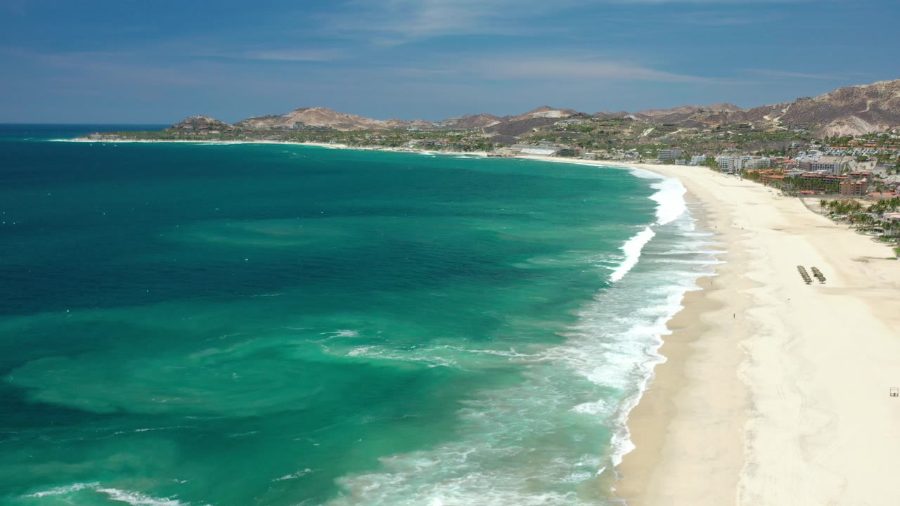 Aerial beach scene with green-blue water, white waves, pale sand, and distant hills.