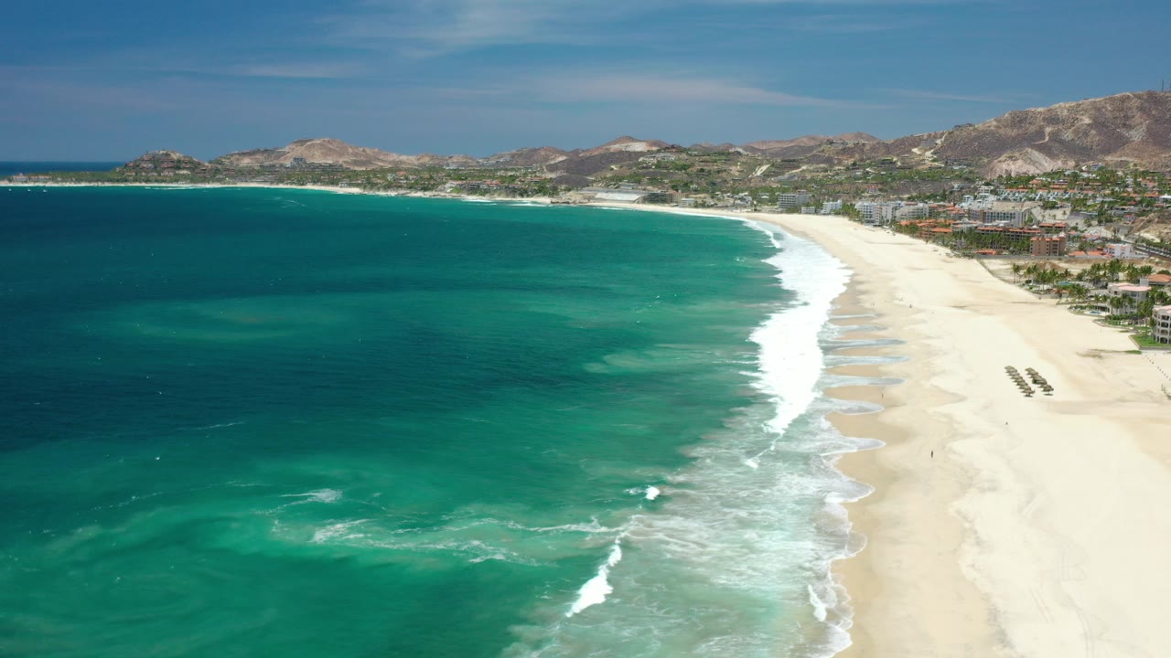 Opening aerial frame over a Los Cabos beach, with turquoise water filling the left side, a pale sand shoreline on the right, distant coastal buildings, mountains, blue sky, and no visible people.