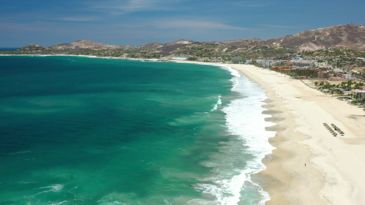 Aerial view of the Los Cabos coastline with turquoise water on the left, white surf curling onto a broad pale beach, low buildings and dry hills in the background, and no visible people.