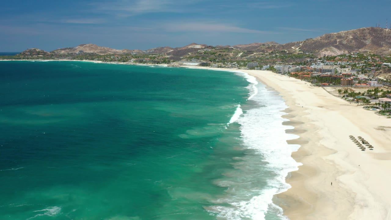 Drone-like view of blue-green water and a white surf band along a Los Cabos beach.