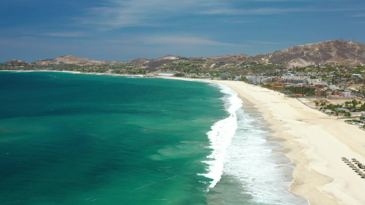 Aerial view in Los Cabos, Mexico with turquoise water on the left, white surf curling onto pale sand, hills on the horizon, and no visible people.