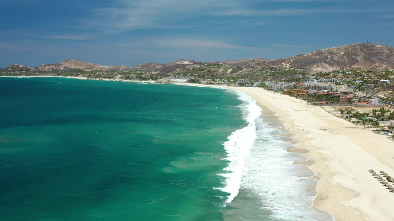 Wide aerial frame over Los Cabos, Mexico showing darker offshore water, bright surf meeting the sand, coastal development to the right, and no visible people.